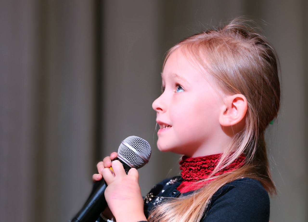 Cute young girl passionately singing into a microphone on stage, captured in a close-up profile view.